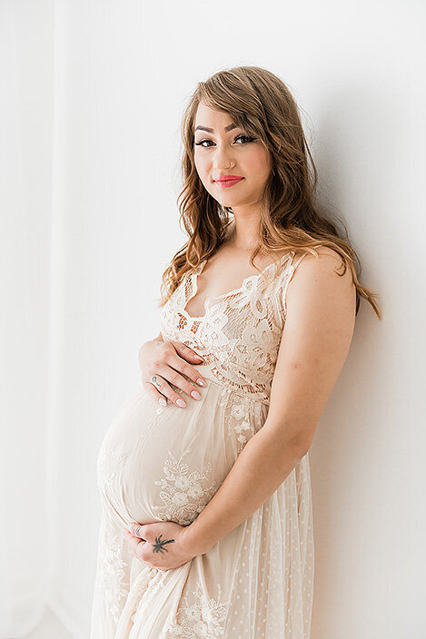 Glowing mother-to-be poses in a sheer embroidered dress against a white studio wall with a calm, confident expression.