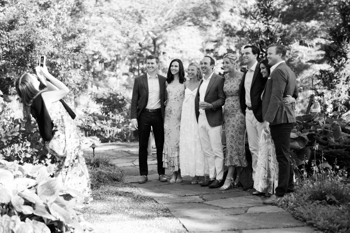 A group of eight friends poses for a photo in a garden setting, while a woman in a floral dress captures the moment with her phone.