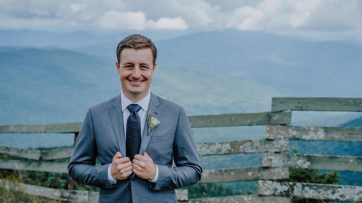 a groom posing in front of a fence and mountains at jay peak
