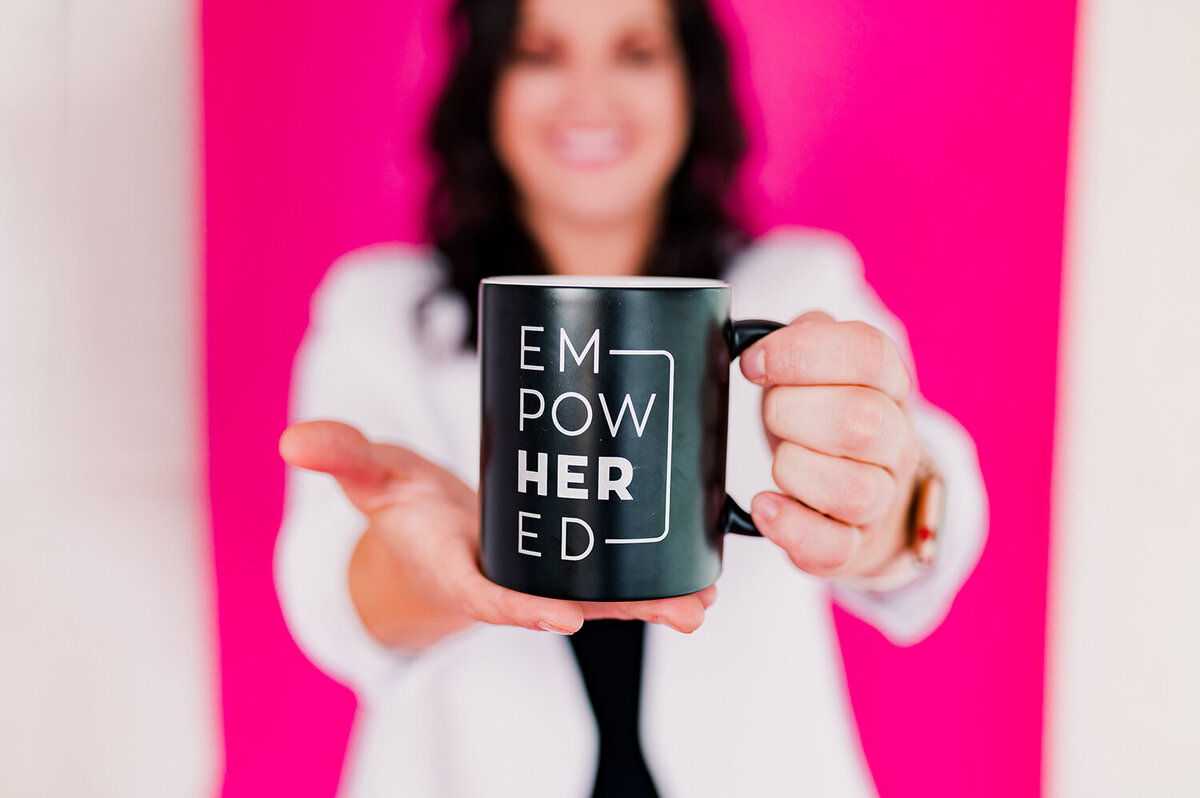 woman in white blazer holding up a black mug that reads "empowHERed"