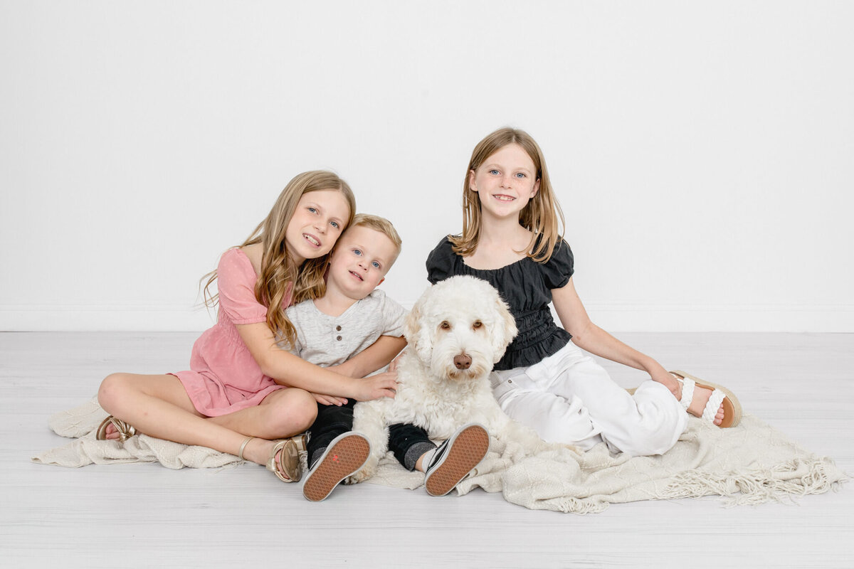 Three young kids sitting with their white, fluffy dog on a cream colored blanket, hugging each other and looking at the camera.