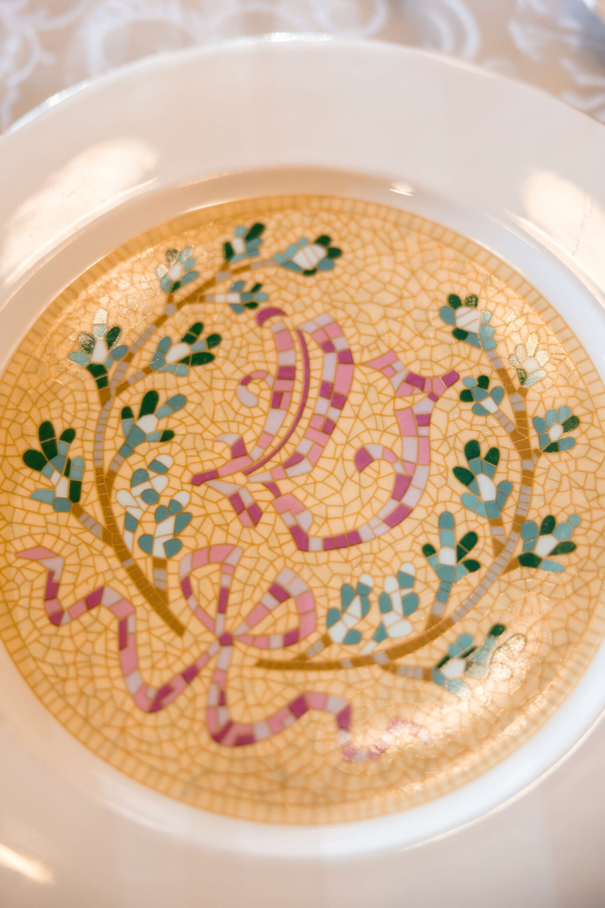 A decorative plate features a mosaic-style design with pastel ribbons, leaves, and floral elements in green and pink tones. The pattern is set against a light, textured background.