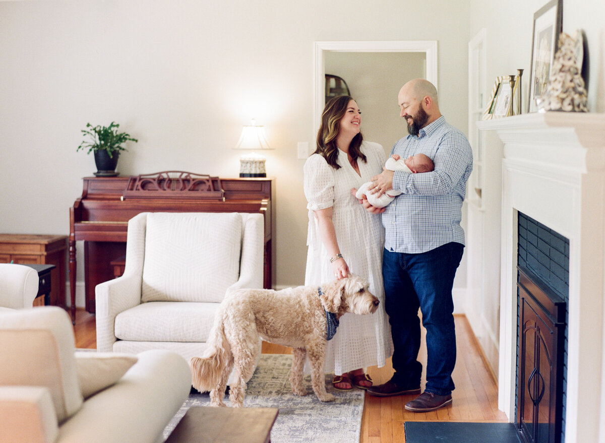 Family standing in their living room, smiling as they hold their newborn son during a Raleigh newborn photography session. Photographed by Raleigh newborn photographer A.J. Dunlap Photography.