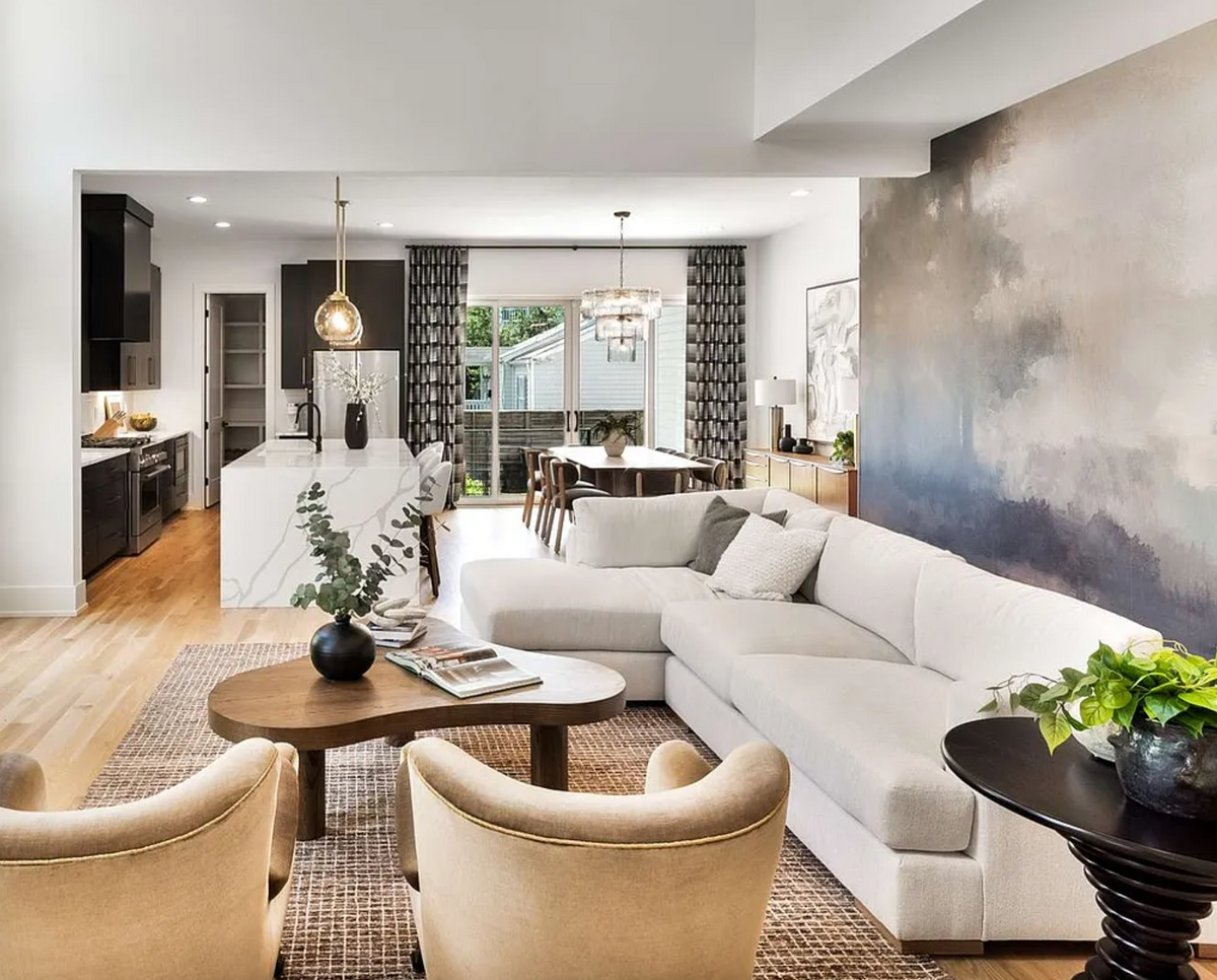 Bright open-concept living room with white sectional, misty forest mural, sculptural coffee table, and modern kitchen with quartz waterfall island