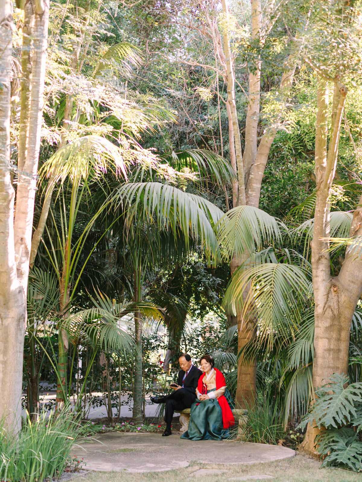 A Korean couple sits on a stone bench in a tranquil, sun-dappled garden surrounded by tall trees and lush greenery.