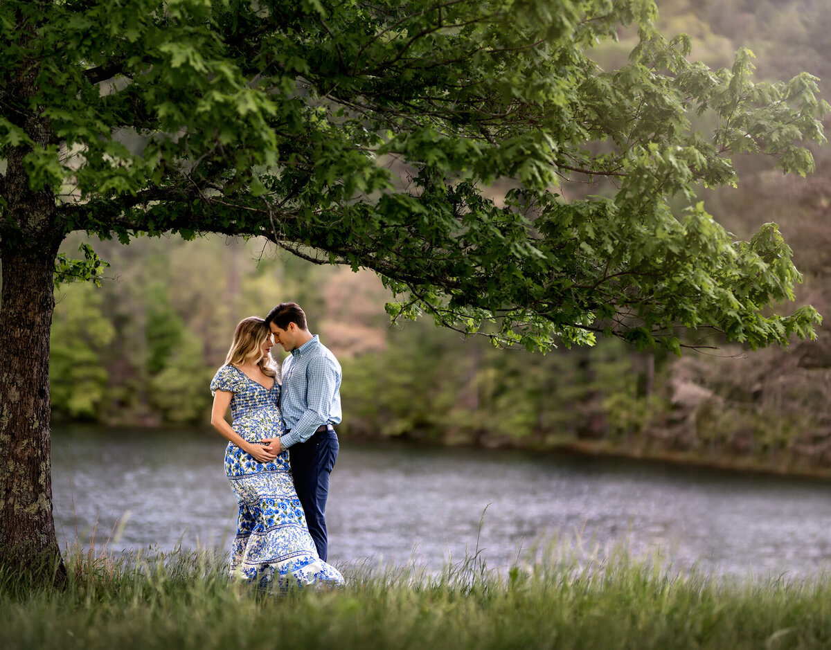 A beautiful couple stands in the tall grass by a lake with  a large tree overhead