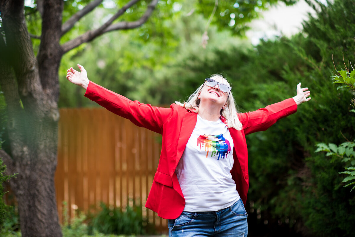 Christine DeHerrera in a red blazer and white t-shirt with rainbow lops, arms thrown side, smiling, outside by a tree with green bushes in the background