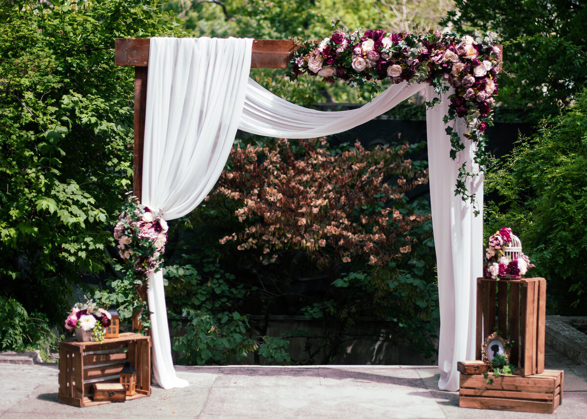 Wedding ceremony arch backdrop decorated with flowers and white drape outdoors.