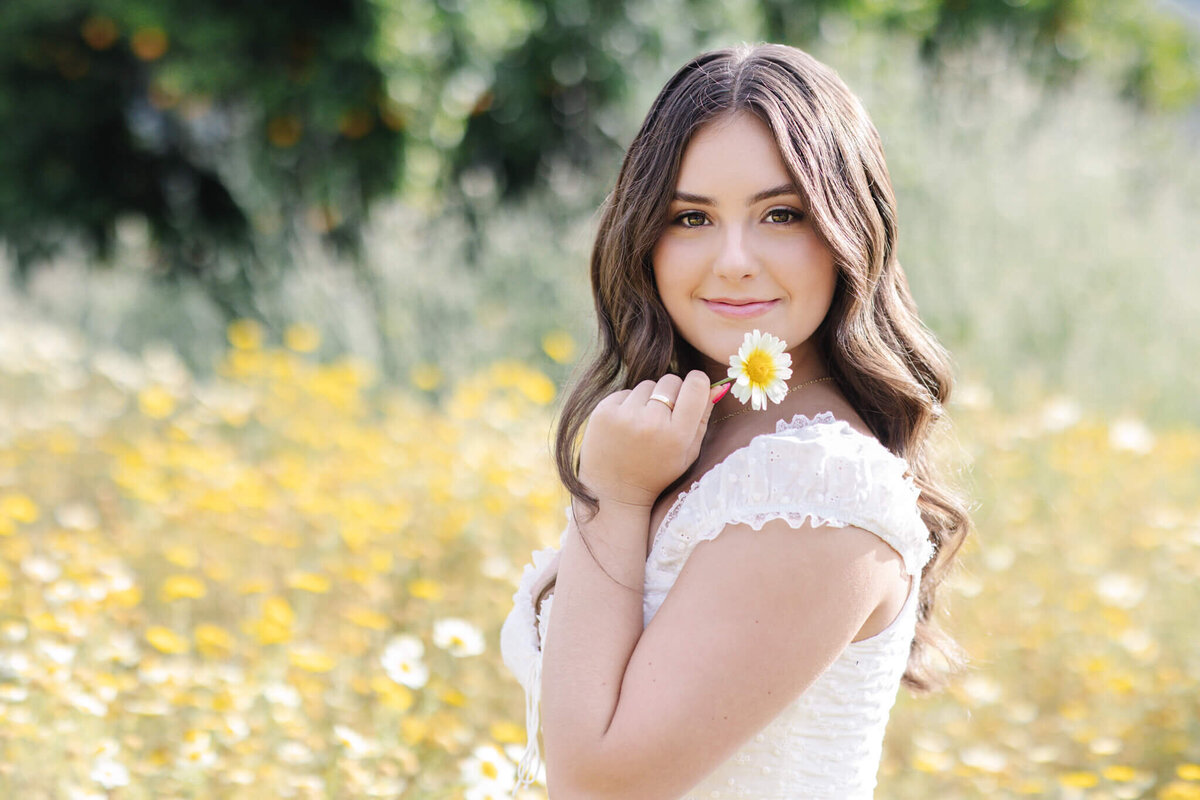 A woman in a white dress holds a daisy by her face, smiling gently. She stands in a field of yellow and white flowers, with greenery blurred in the background.