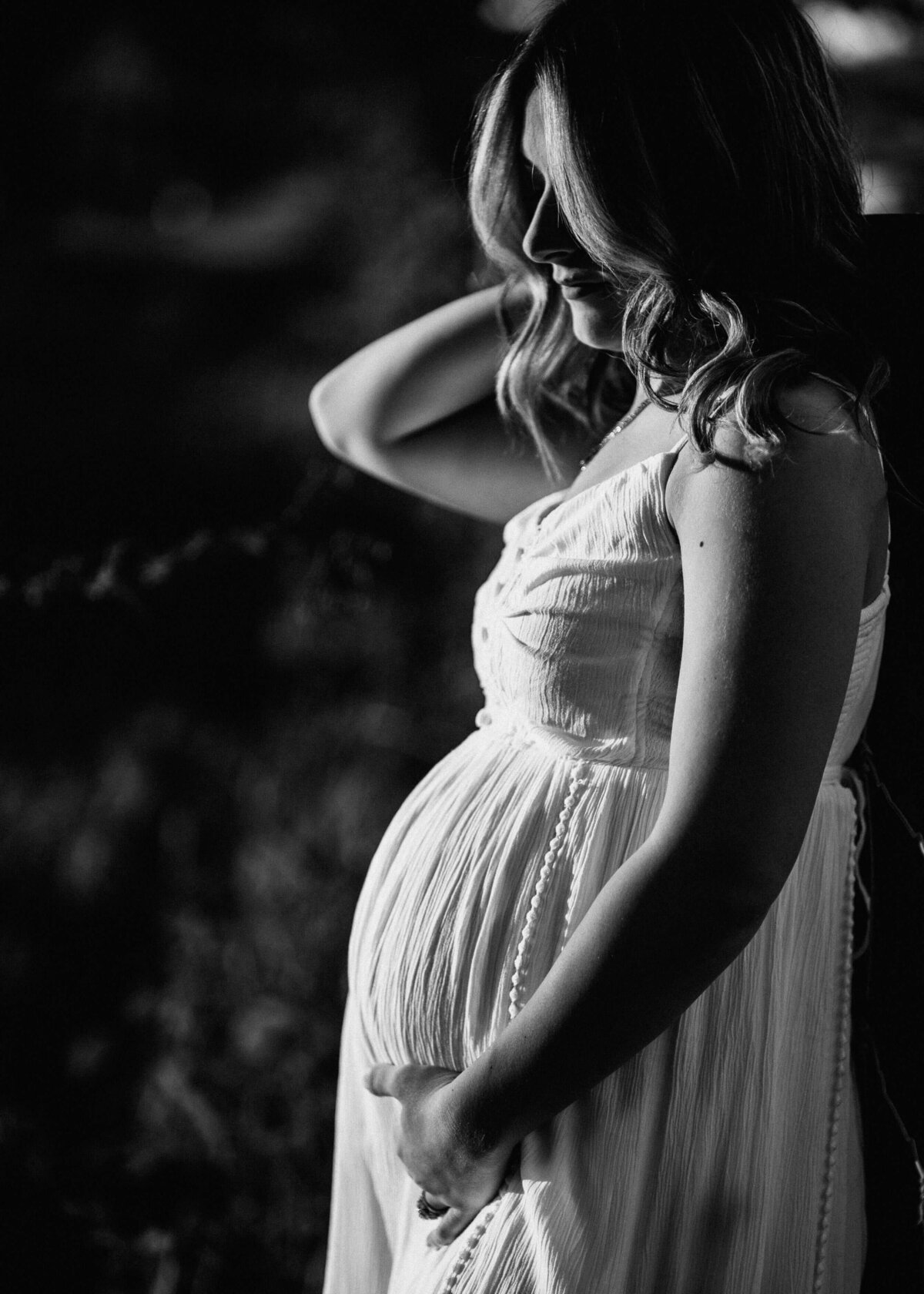 A pregnant woman in a white dress leans against a fence, captured beautifully by a Pittsburgh maternity photographer.