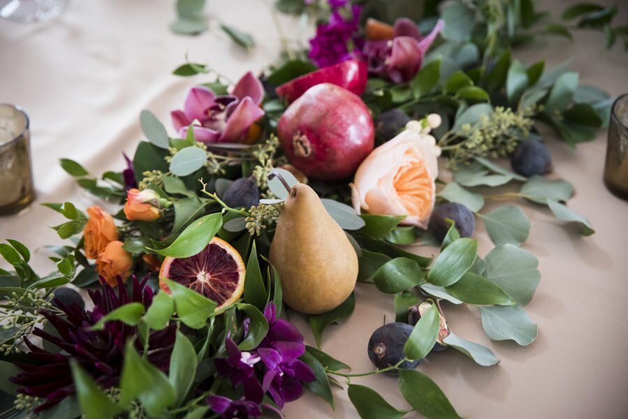 A close up shot of table greenery featuring purple and maroon florals with pears, fig and other fruit.