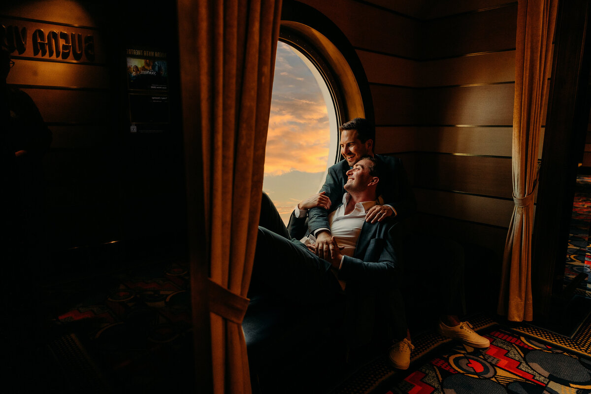 One groom lounges in the other’s arms near a round Disney Fantasy porthole with glowing sunset views behind them.