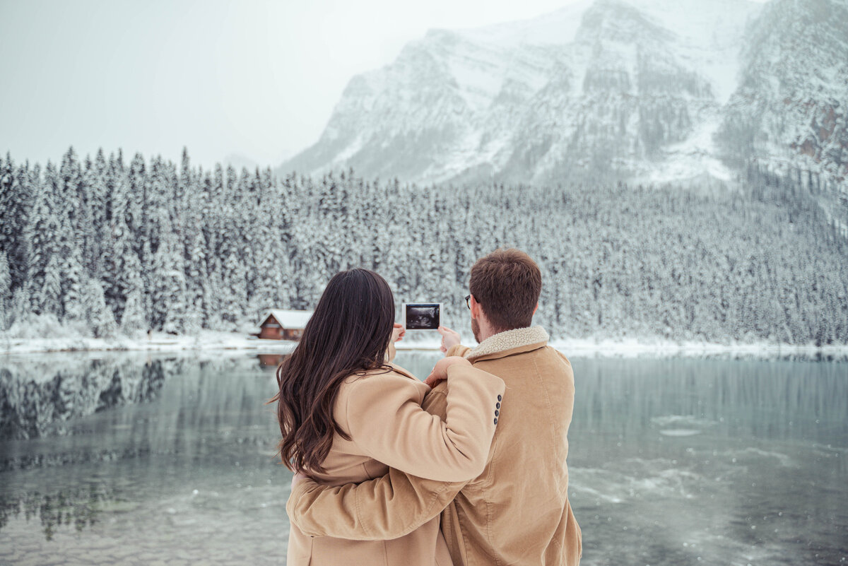 Mother and father holding up their sonogram while looking at a winter Lake Louise.