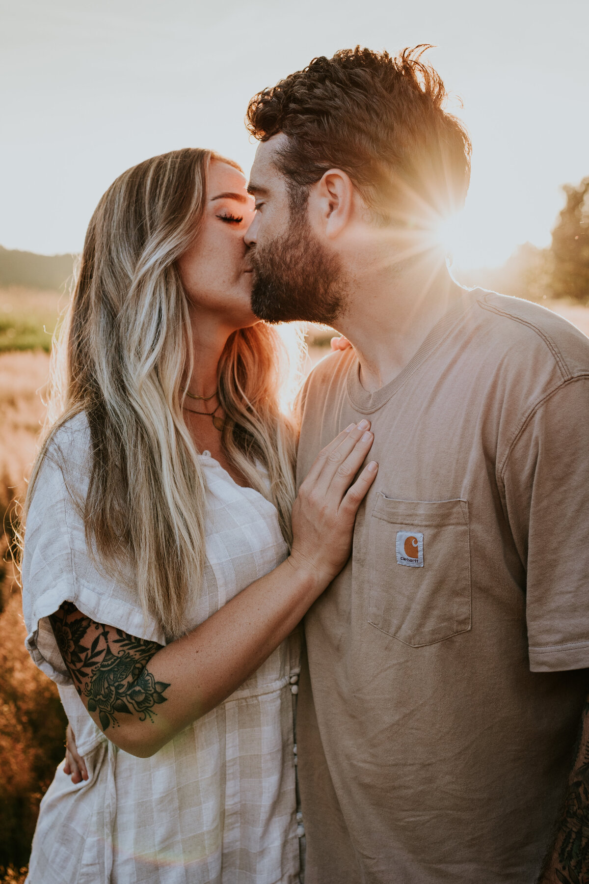 Man kisses wife in open field while sun sets behind them.