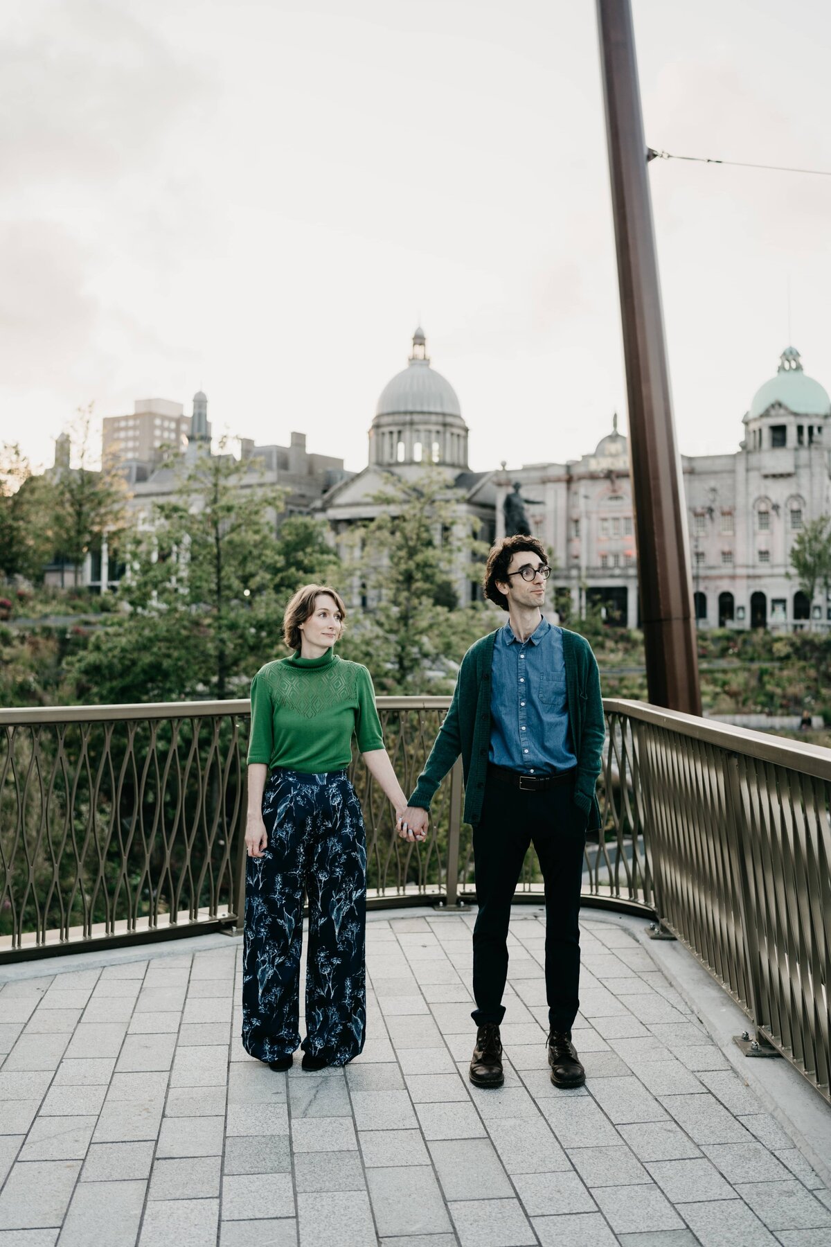 A alternative couple hold hands in Union Terrace Gardens during their enagagement photoshoot. They stand with HIs Majesty's Theatre in the background.