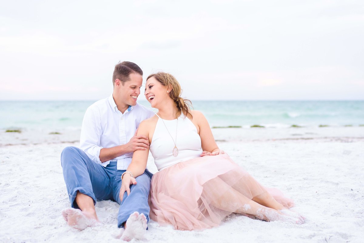 Soon to be bride and groom laughing while sitting on the white sandy beaches of Tampa Bay