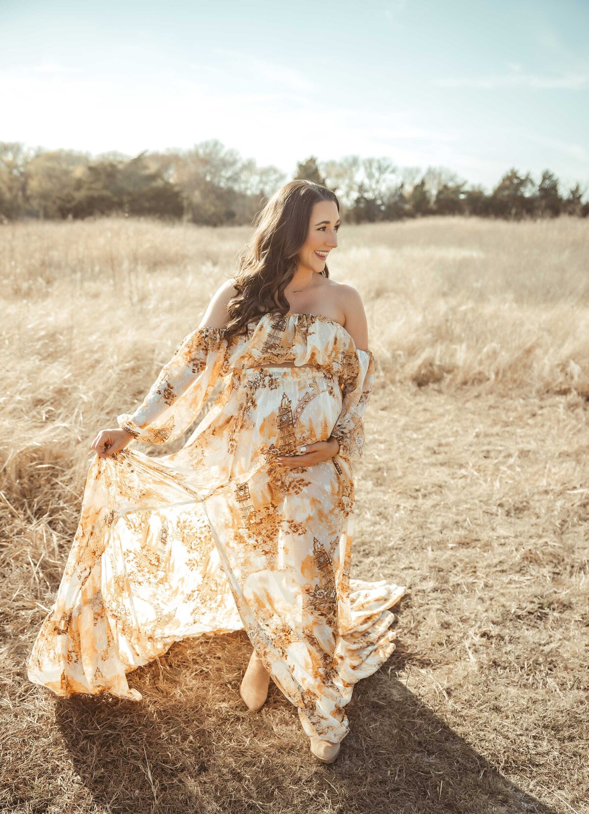 Maternity photo of a young woman walking in the sun light moving her flowy dress with one hand and holding her stomach wit the other hand
