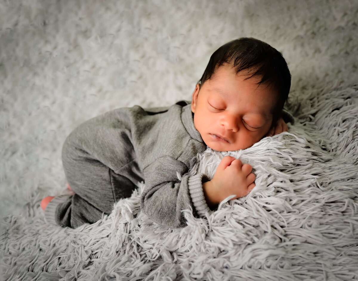 Image by Beth Benecki of E. Benecki Photography of a newborn in a gray outfit sleeping peacefully on his belly  on a gray fur cover.