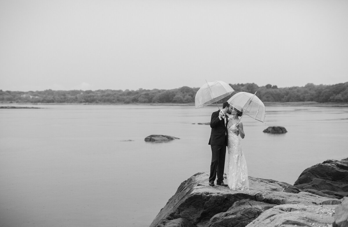 Bride and Groom with umbrellas pose by the water during rainy newport wedding day.
