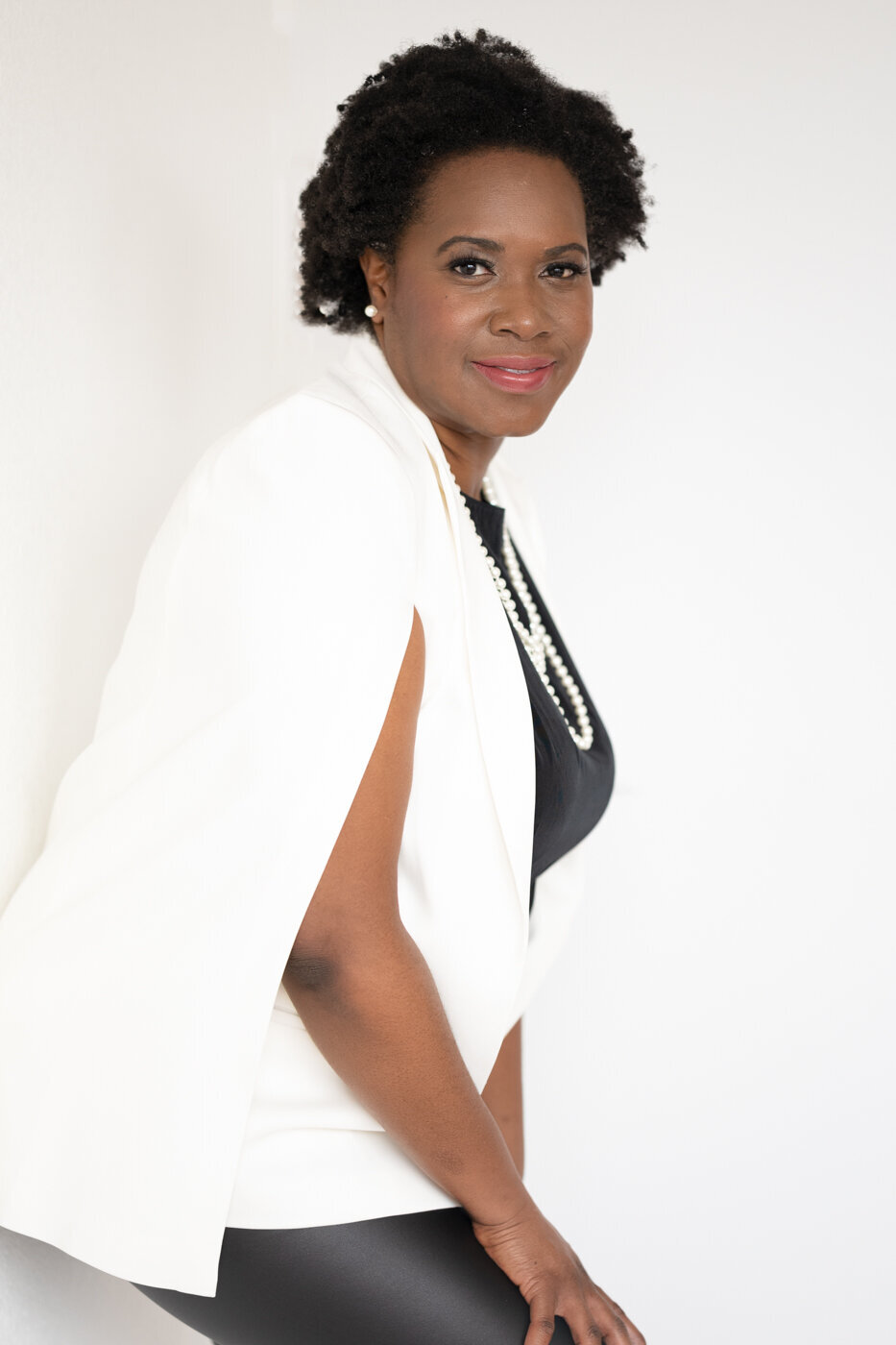 A black woman with short curly hair posing on a white wall wearing a black outfit with a white jacket touching her leg with one hand