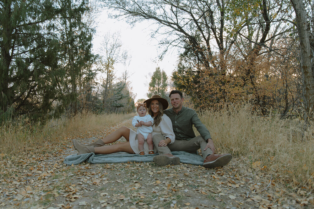 A husband and wife sit on a blanket while holding their baby girl on their lap. They're smiling for a posed family photo.