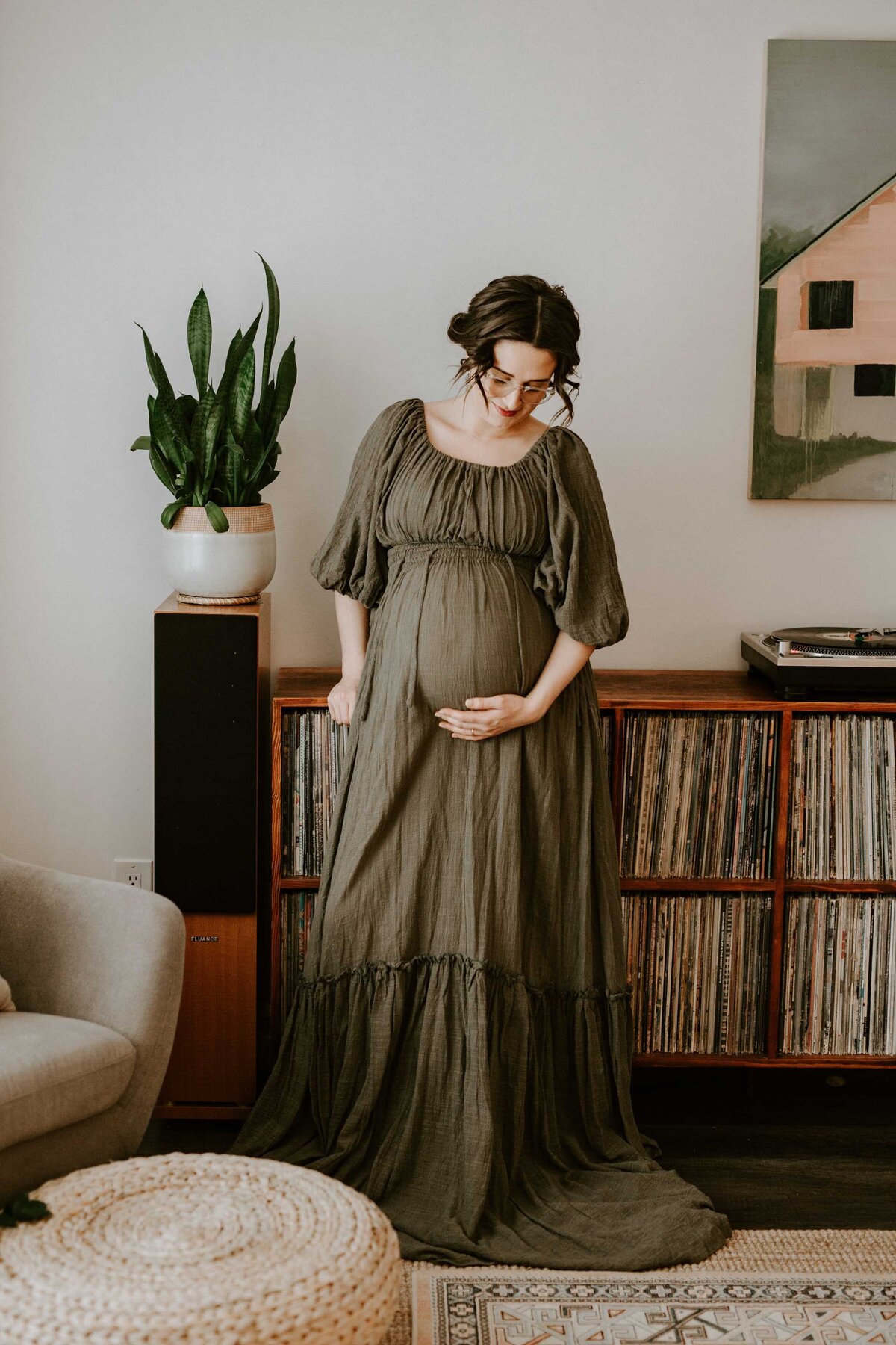 In home lifestyle maternity session. Expectant mom in long green maternity gown is looking at and touching her baby bump while leaning against shelves that house a large record collection.