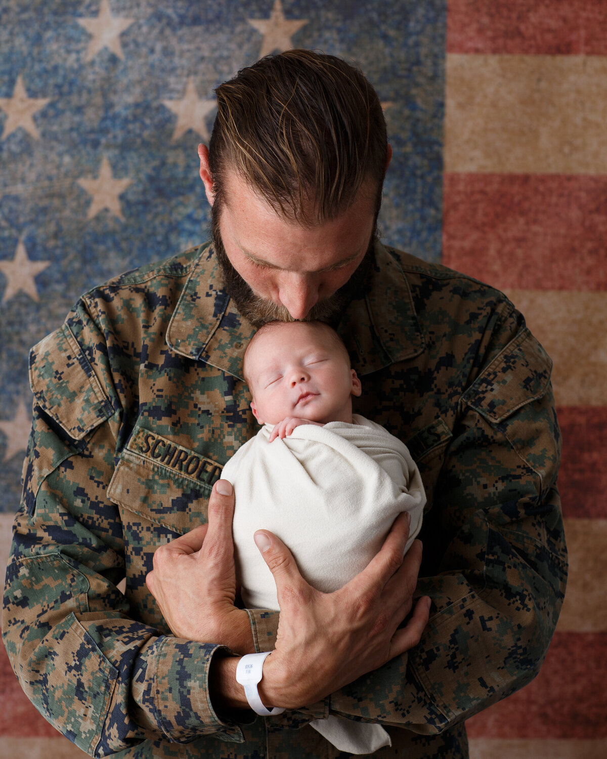 Father wearing military fatigues holding his newborn son in his arms and photographed on an american flag background by newborn photographer at Life in Pink Photography