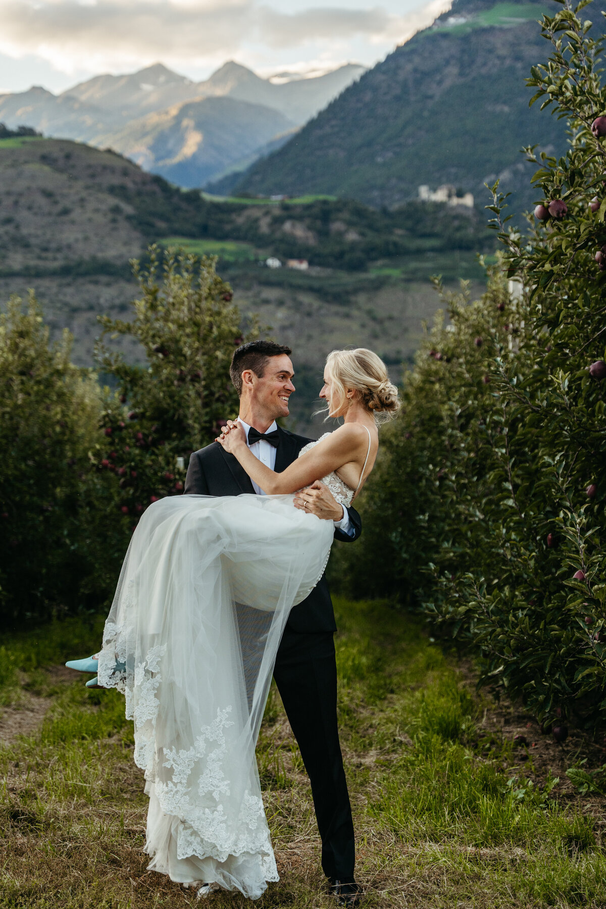 Groom lifting bride in orchard with mountain backdrop