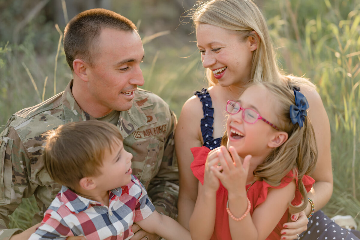 Family wearing military uniform and patriotic clothing laughs together in tall grasses.