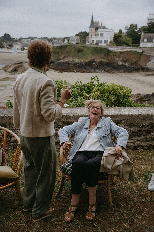 Une invitée âgée rit aux éclats sur la plage de Saint-Quay-Portrieux. Un instant spontané capturé par Kamescop, photographe et vidéaste de mariage.
