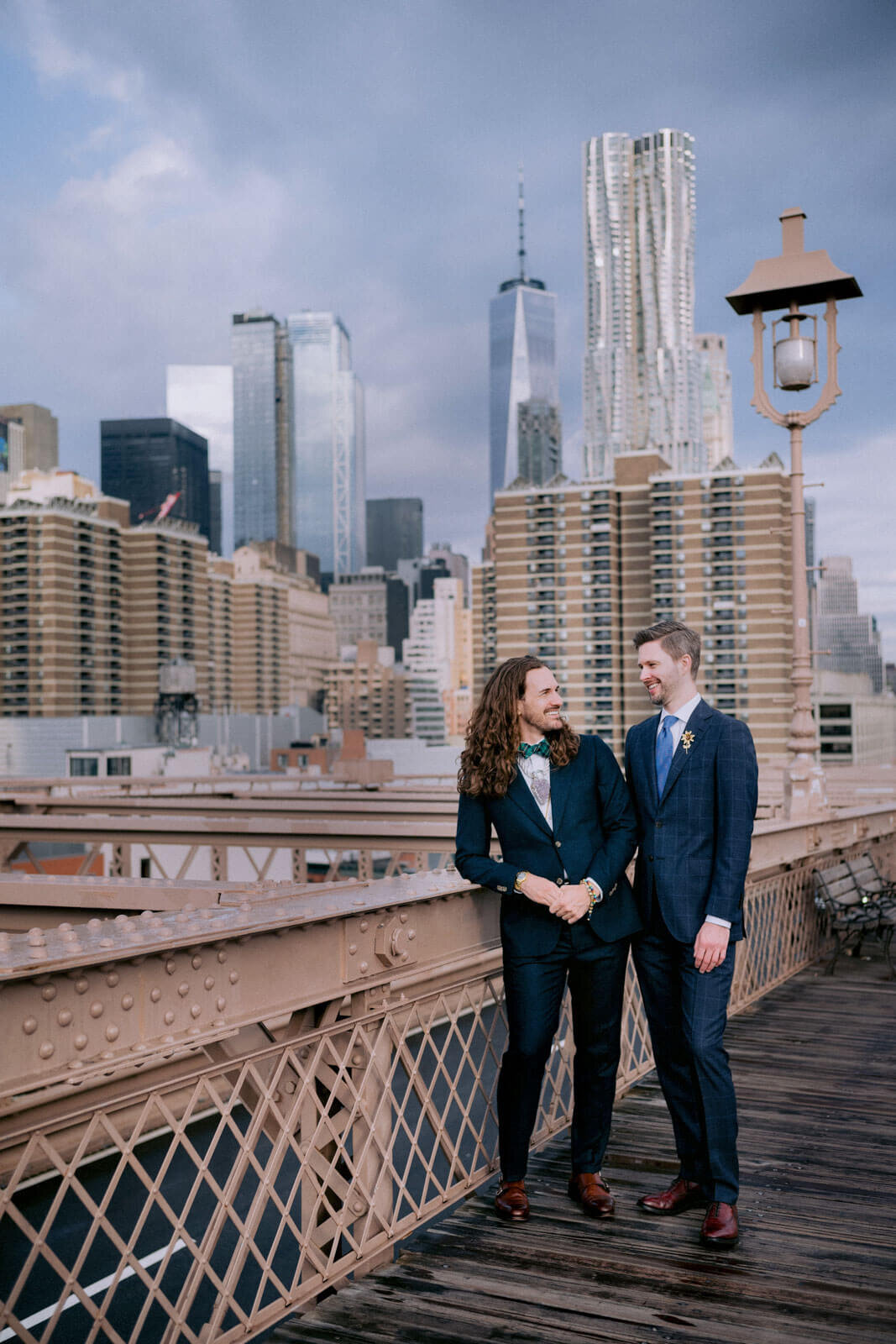 The two grooms are chatting on the Brooklyn Bridge, with skyscrapers in the background. NYC Elopement Image by Jenny Fu Studio