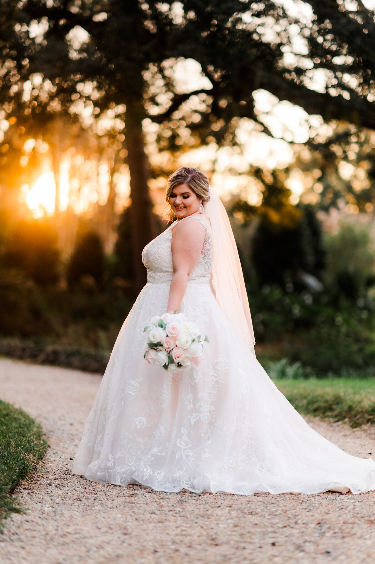 sunset bridals with Little Rock wedding photographer as the bride walks down a gravel path in her lace wedding dress as she looks down her arm to her white peony wedding bouquet