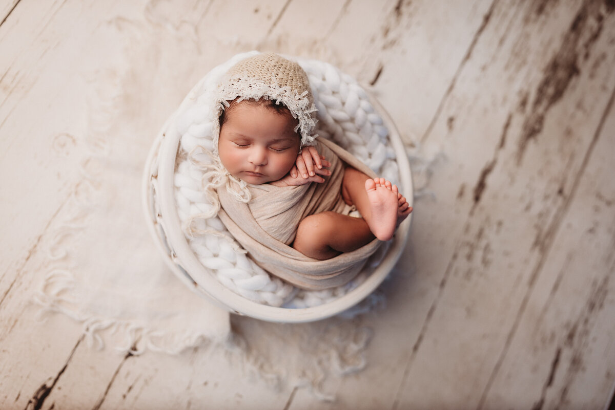 2 week old baby girl wrapped in beig wrap wearing baby bonnet wrapped  in white basket bowl prop on wooden floor drop
