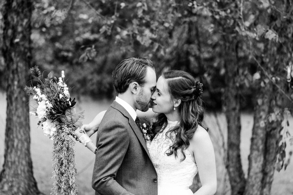 Bride and groom sharing a kiss in a wooded area, captured in black and white. The romantic setting among the trees enhances the classic and elegant atmosphere of their wedding day.