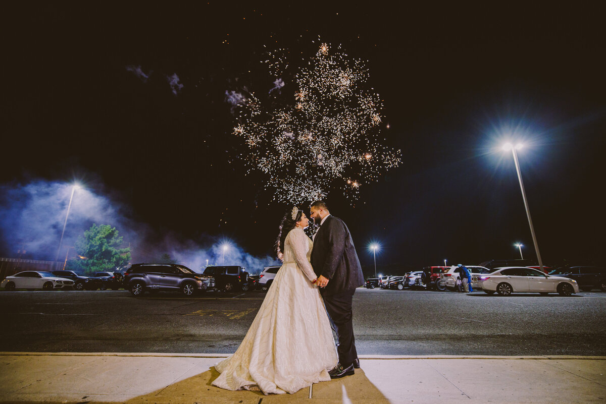 Indian wedding couple share a moment in the reception parking lot with fireworks in the night sky