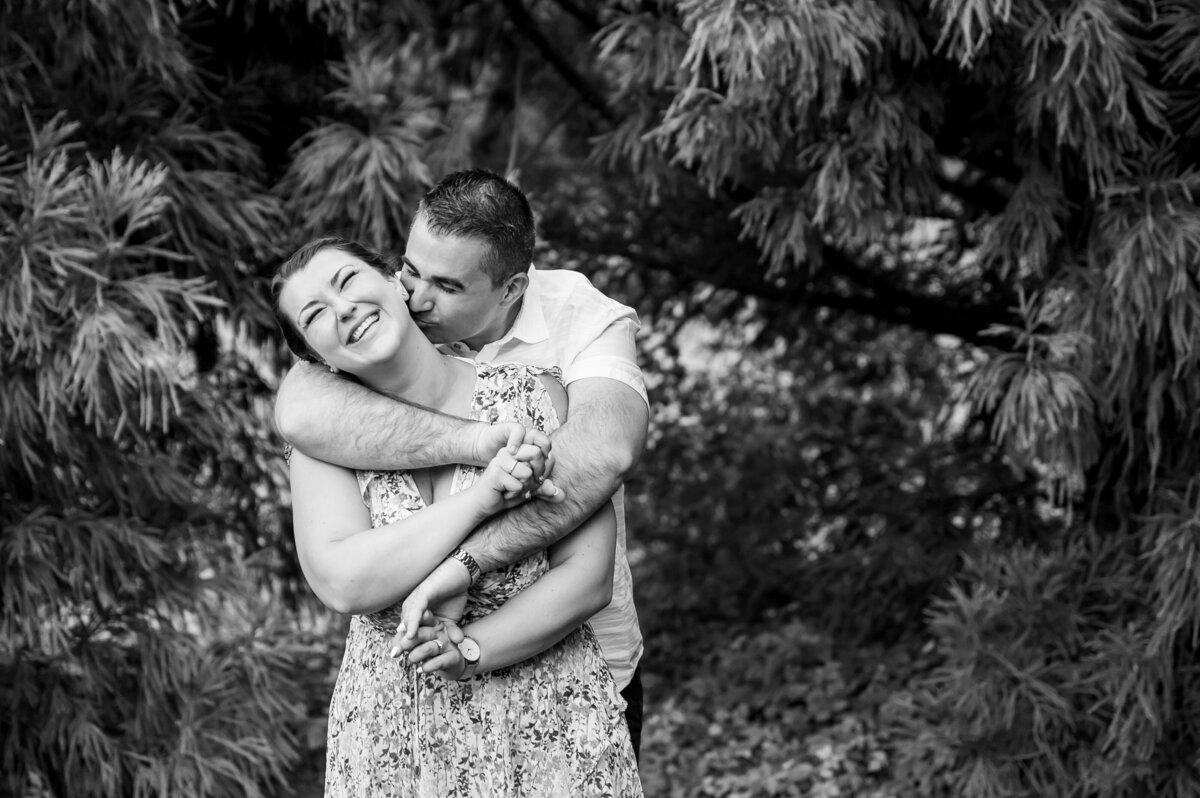 A couple stands in a wooded area, with the man playfully embracing the smiling woman from behind. They are surrounded by trees and foliage. Black and white image.