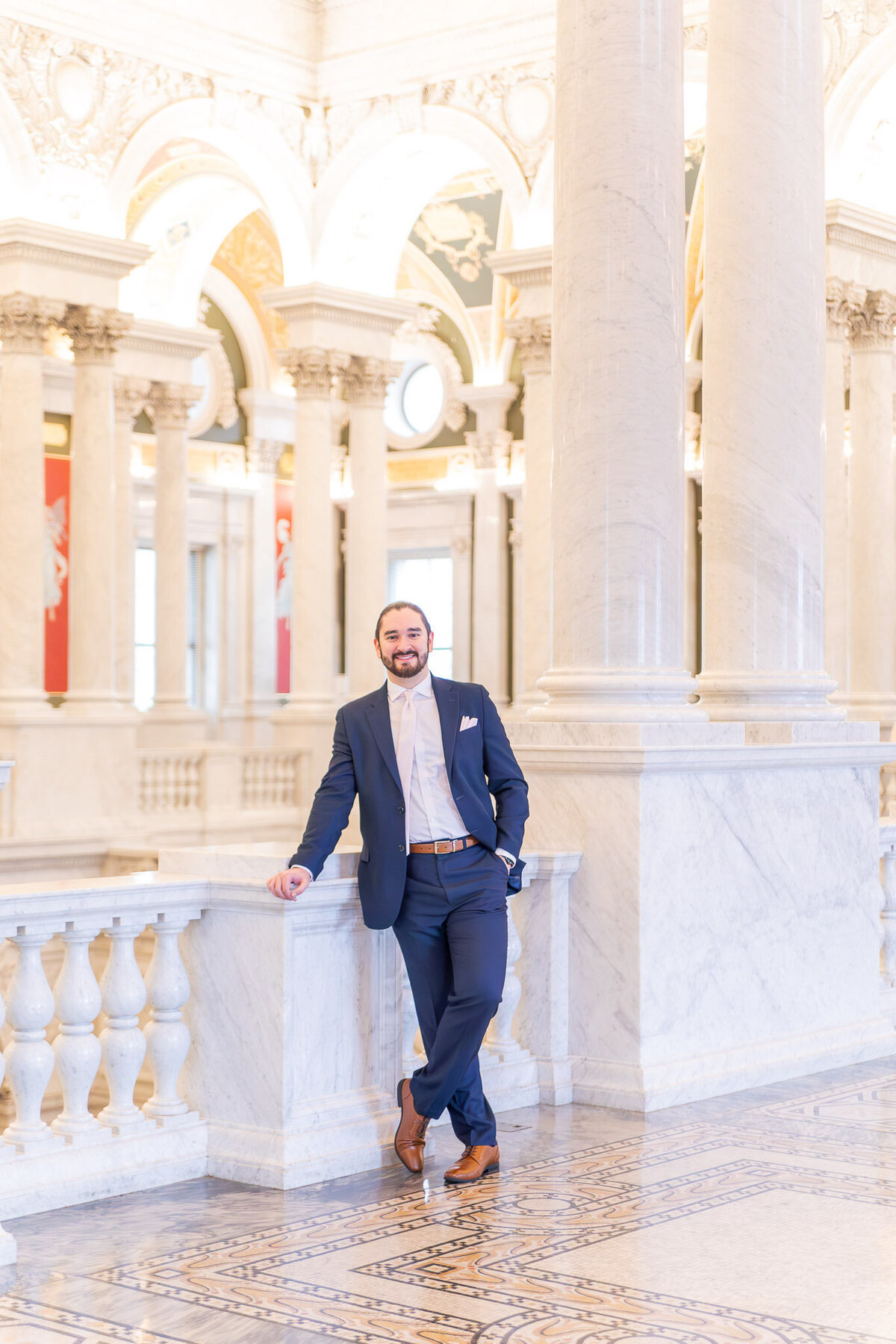 A man poses for a portrait inside of a fancy building in Washington D.C. He is dressed in a classic blue suit and is smiling at the camera.