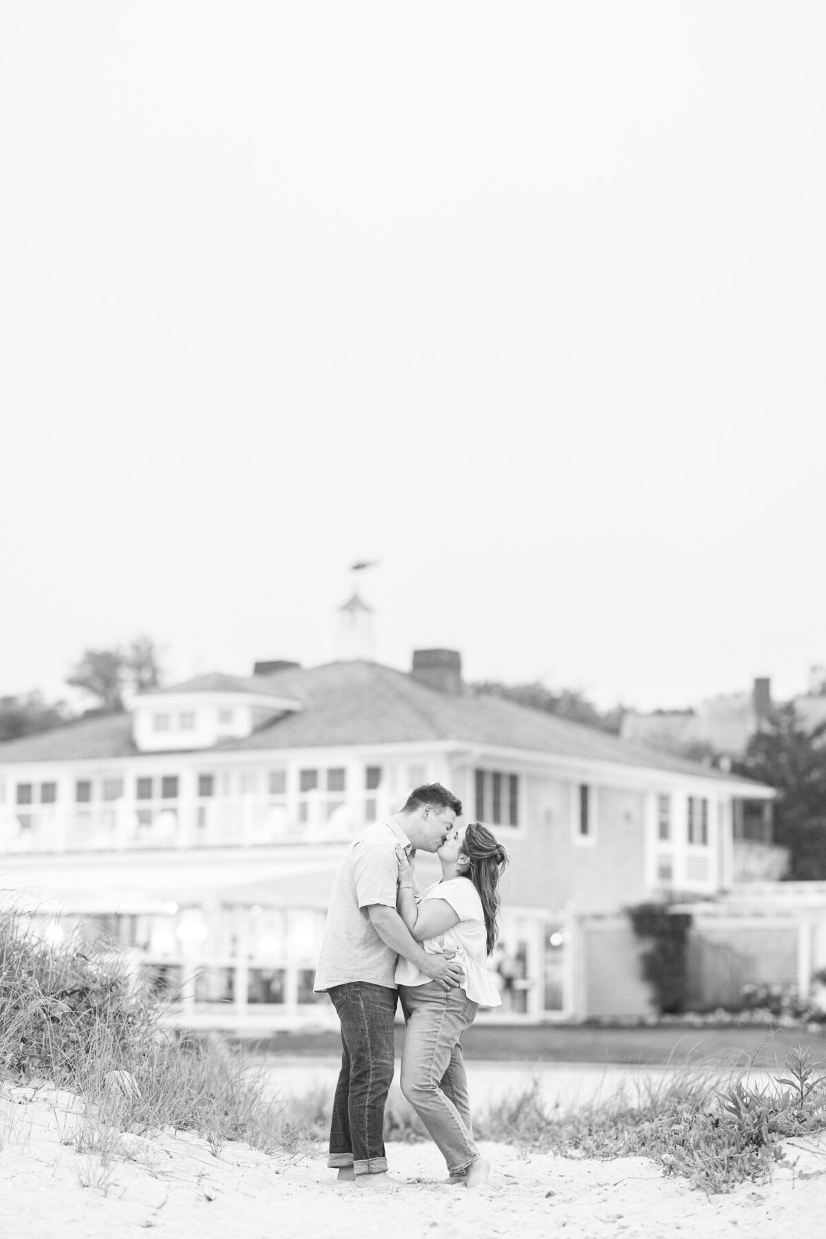 Black-and-white photo of a couple kissing on the sandy beach with Chatham Bars Inn in the background — representing the portfolio of a Cape Cod engagement photographer.