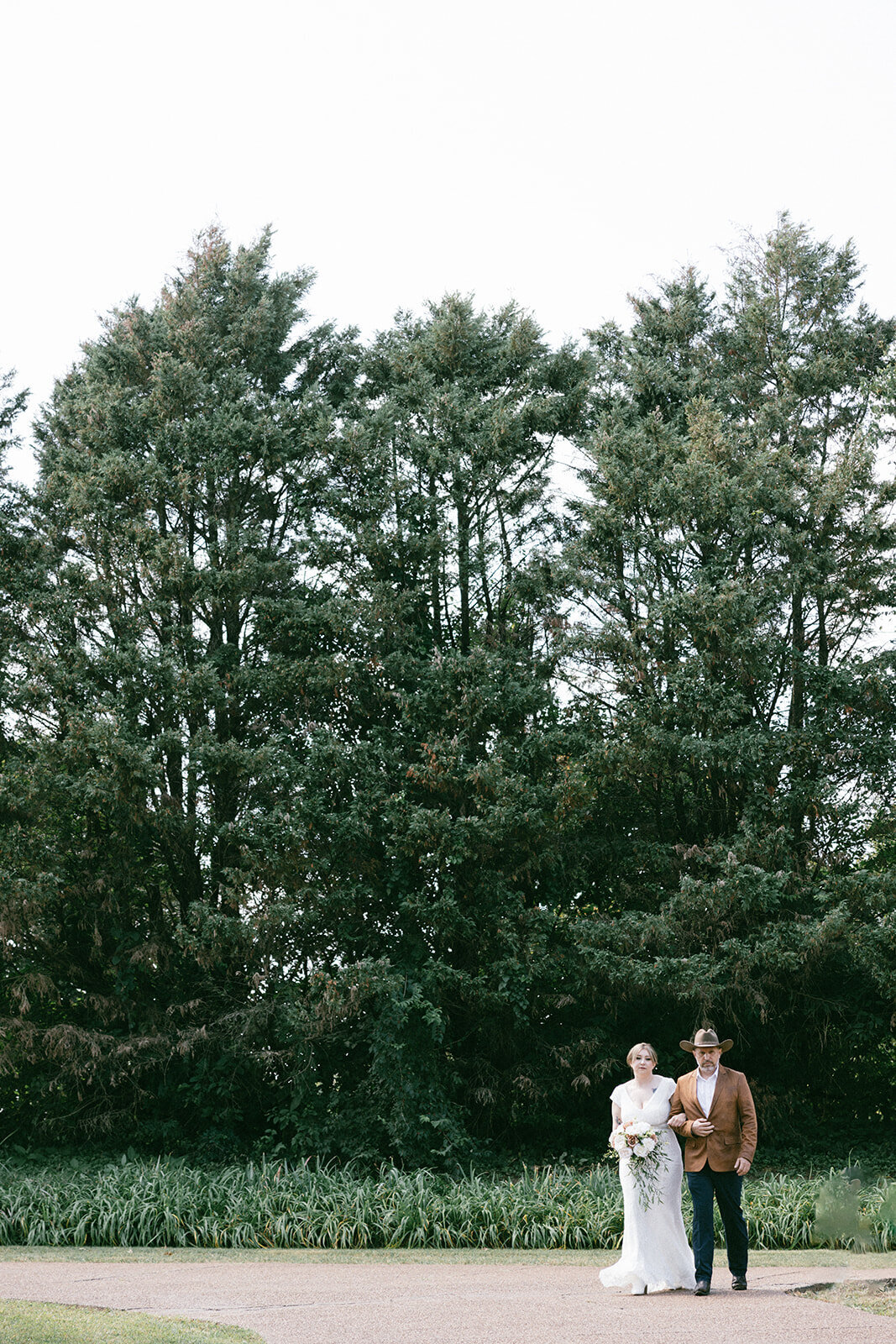 Father in brown cowboy hat walks his daughter in a white wedding gown down the drive to an elopement surrounded by trees. 