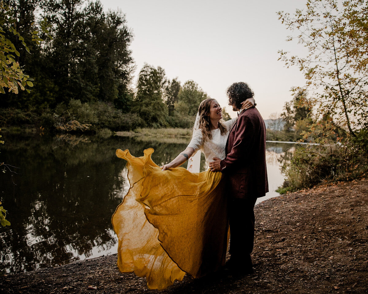 Bride swishes her mustard dress while kissing her groom by a lake