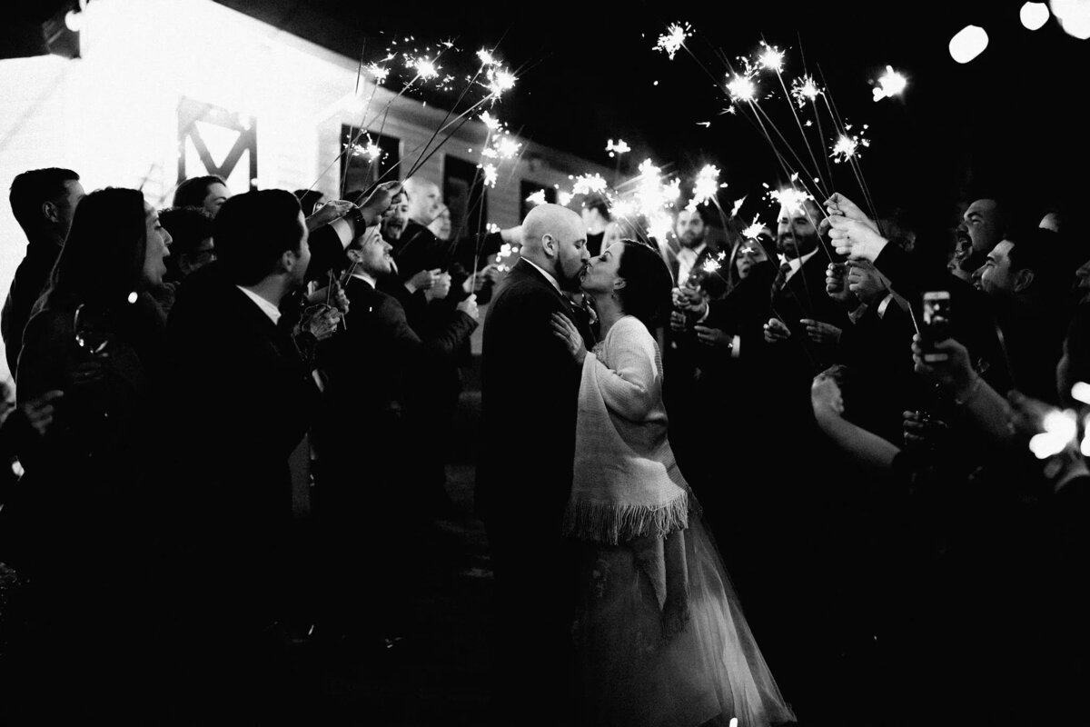 A couple kisses surrounded by a crowd holding lit sparklers at night, creating a festive and romantic atmosphere. Captured by an NJ wedding photographer, the lively scene glows with joy and love.