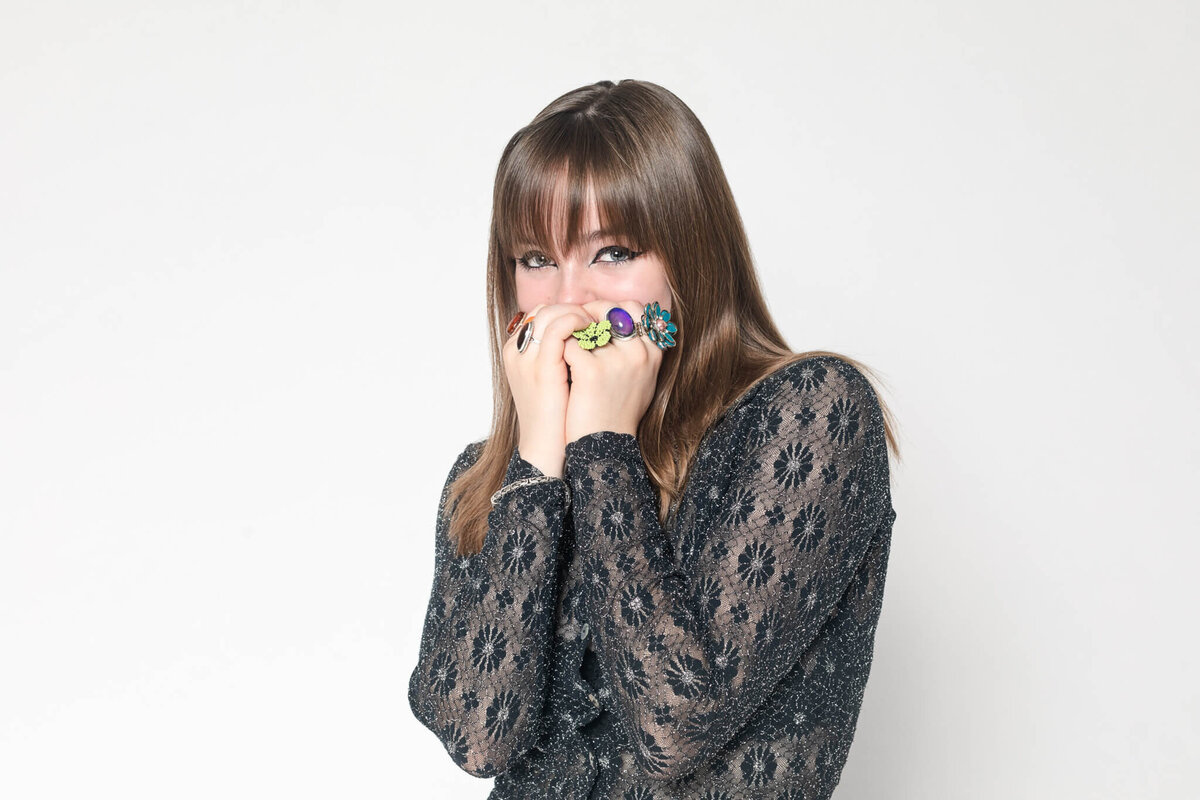 A woman with long brown hair is wearing a black lace top. She is playfully covering her mouth with her hands, which have colorful rings, and looking at the camera. The background is plain white.