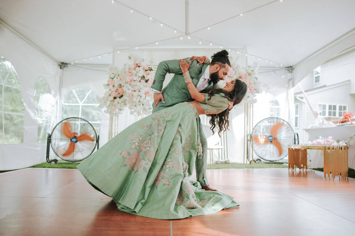 A groom dips his bride gracefully on a dance floor under a decorated tent, her green floral lehenga flowing elegantly.