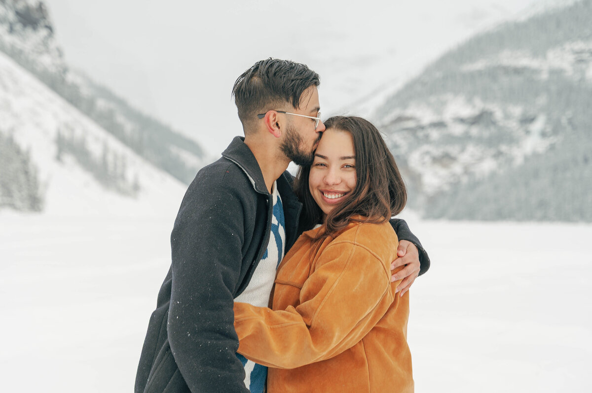 Couple shares a kiss on frozen Lake Louise during winter engagement.