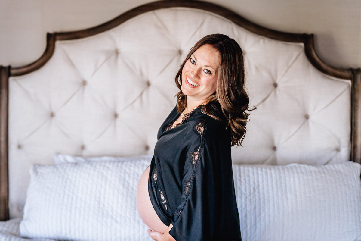 A pregnant woman stands smiling, holding her belly while standing in front of a tufted headboard, her dark robe draped over her shoulders.