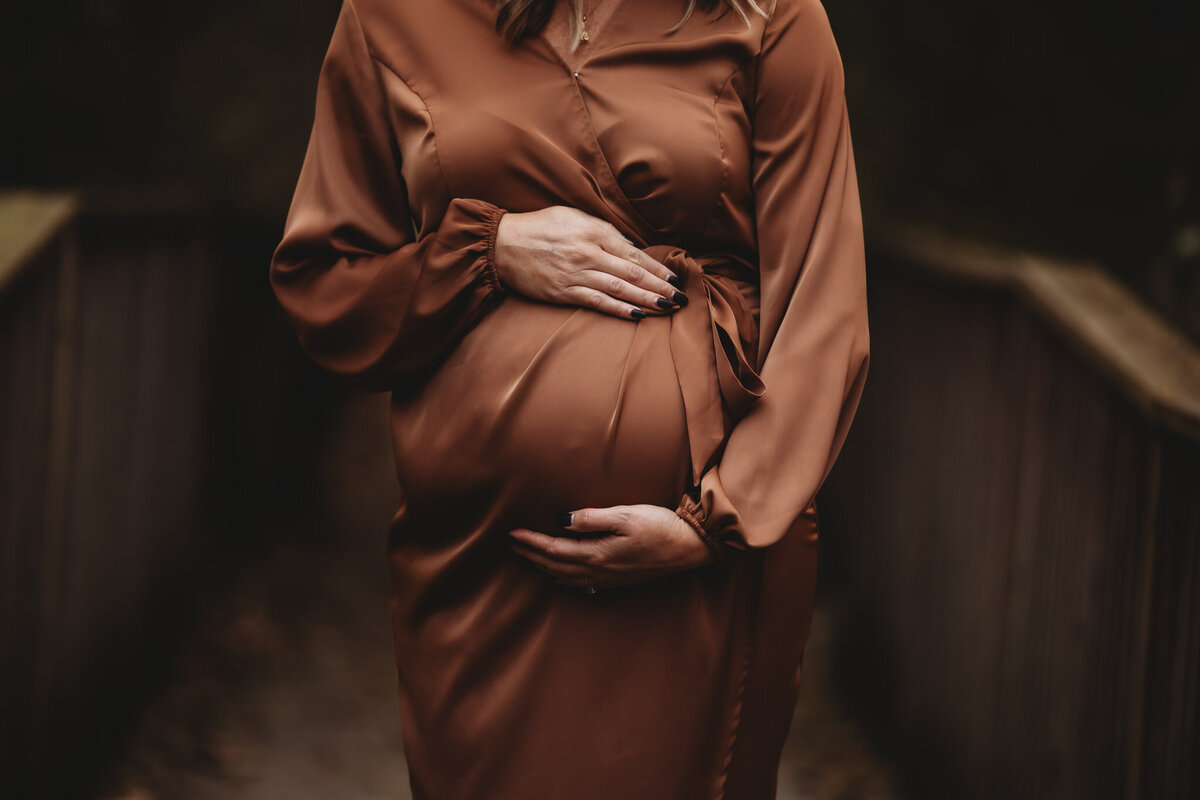 Photo of pregnant woman neck down waist up showcasing her pregnant belly in a warm brown dress outside