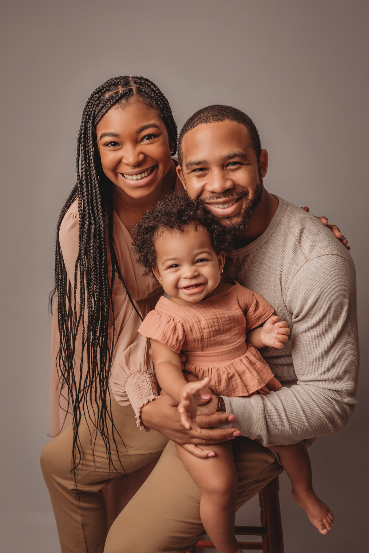 Family portrait of mom, dad and one year old baby girl with baby on dad's lap and mom leaning in behind them and family wearing nude pinks and creams, smiling at the camera