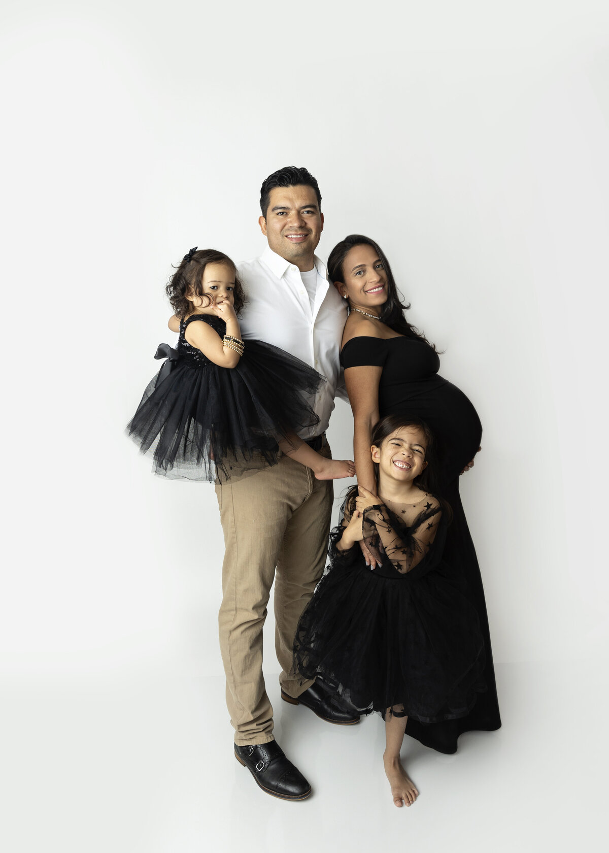 An expecting mother stands in a studio with leaning on her husband with their two toddler daughters in matching black dresses