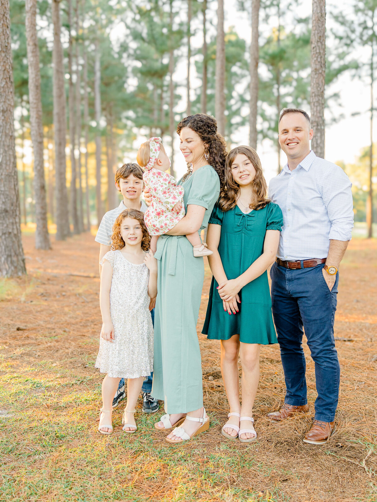A Fayetteville NC family photographer captures a family of six posing outdoors among tall trees, sunlight filtering through. The mother holds a baby while two girls and a boy stand nearby. The father stands next to them, all casually dressed and smiling, in this beautiful moment.