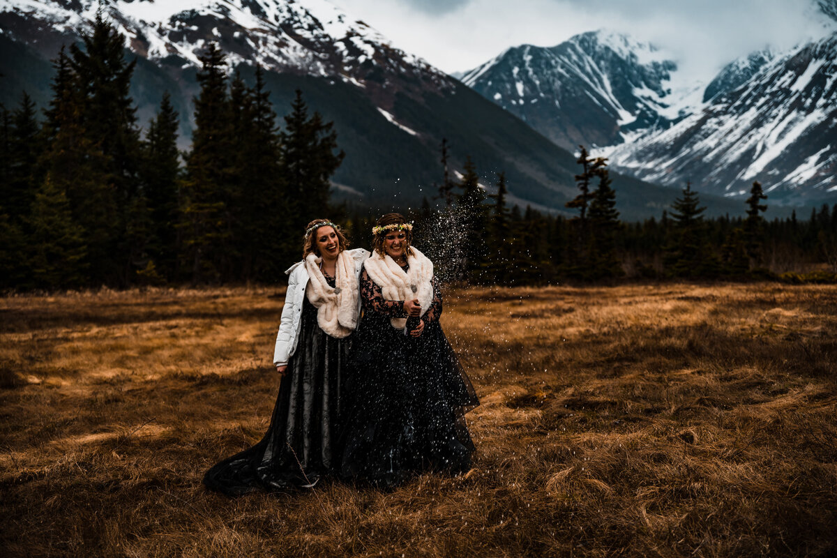 Both brides wearing black wedding gowns and white fur stoles, look super excited after popping champagne following their elopement ceremony in Alaska.